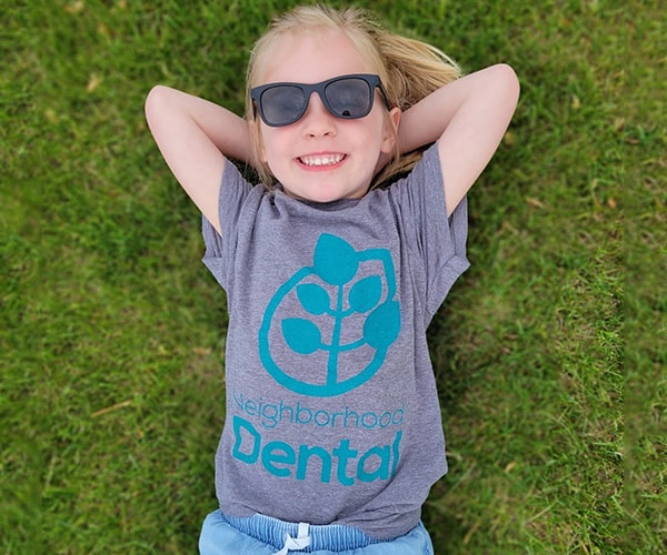A girl patient at Neighborhood Dental lying on the grass while smiling in a shirt from our dental office